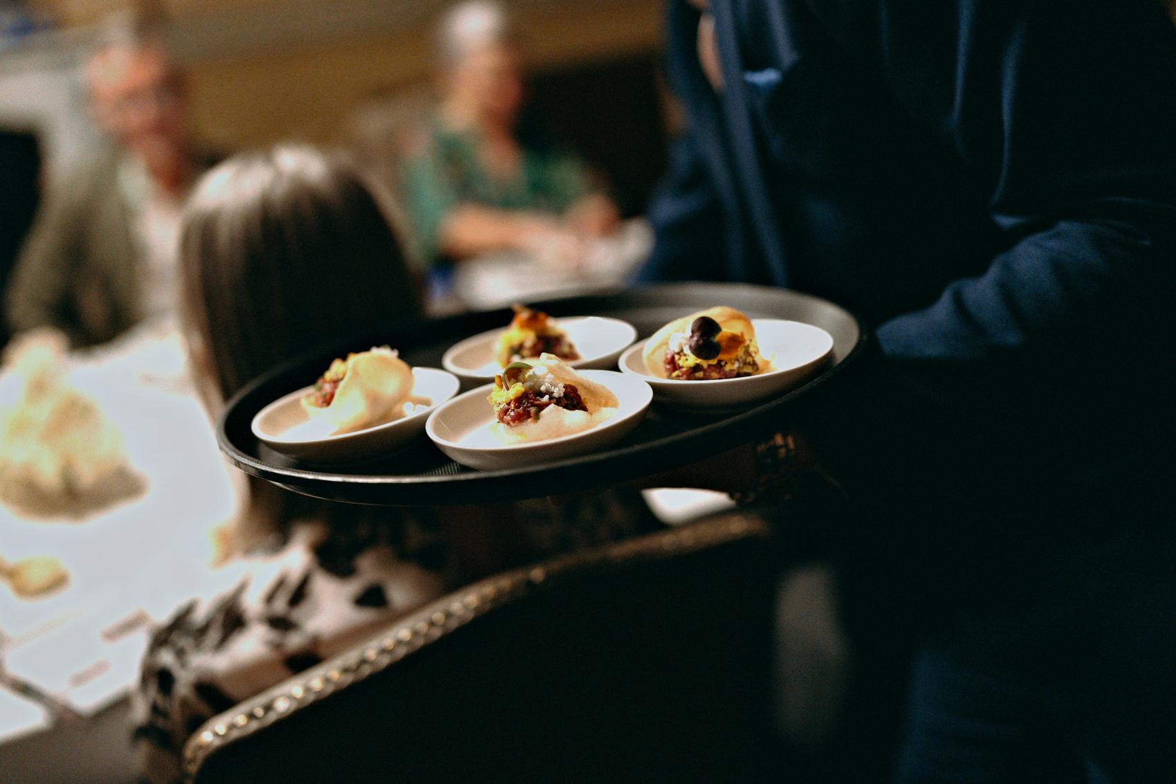A waiter carrying a tray of canapes through a seated room