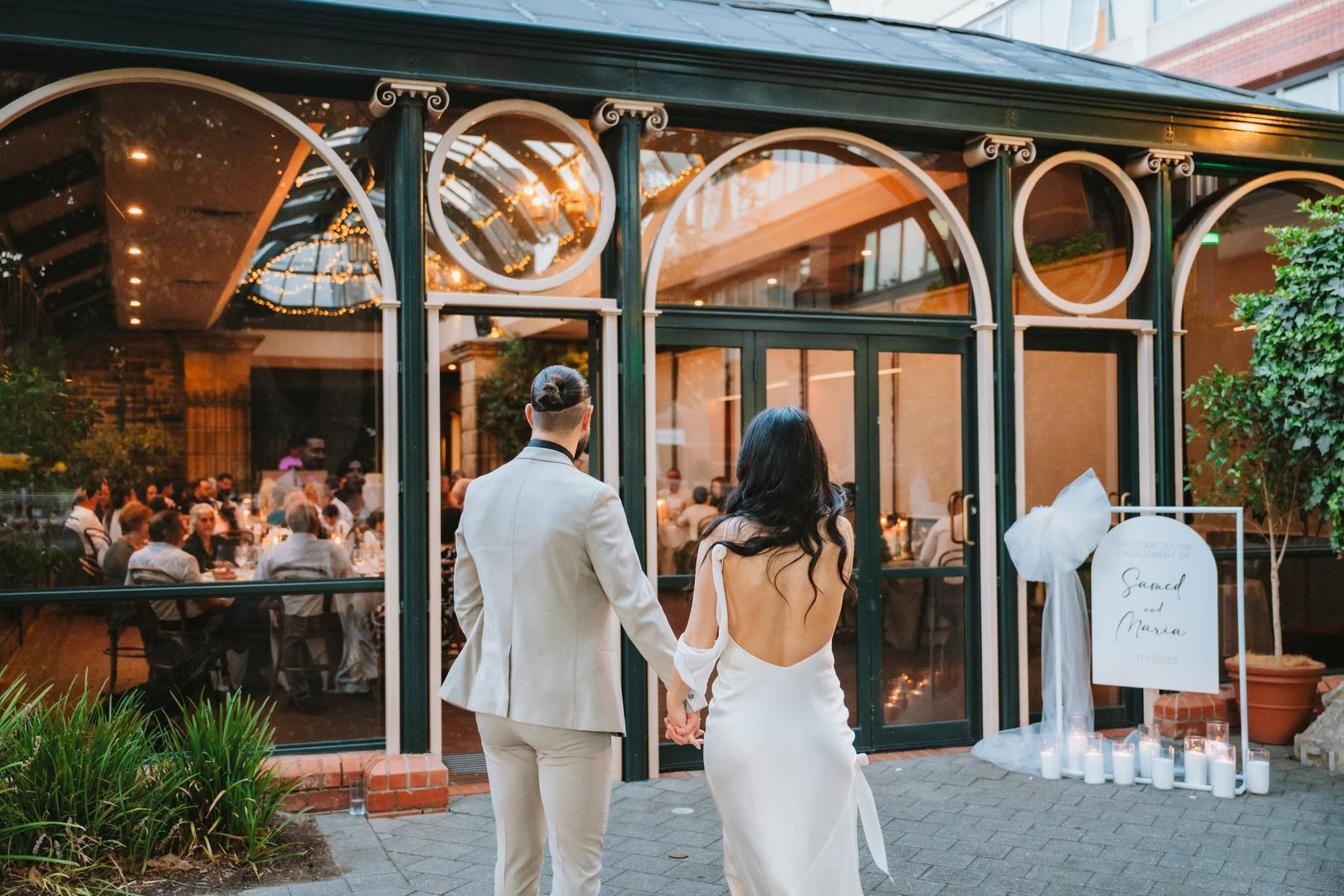 A couple walking up to the lit Conservatory at Ayers House at dusk, guests inside at candlelit tables