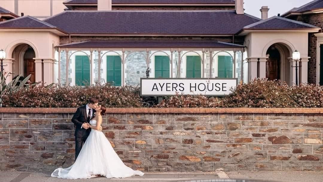 A couple sharing a kiss beside the Ayers House sign on the bluestone wall out the front